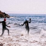 Full body side view of people in wetsuits and swim caps coming into wavy ocean in daytime