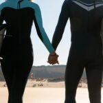 Couple in wetsuits holding hands with surfboards on a sunny beach in Portugal.